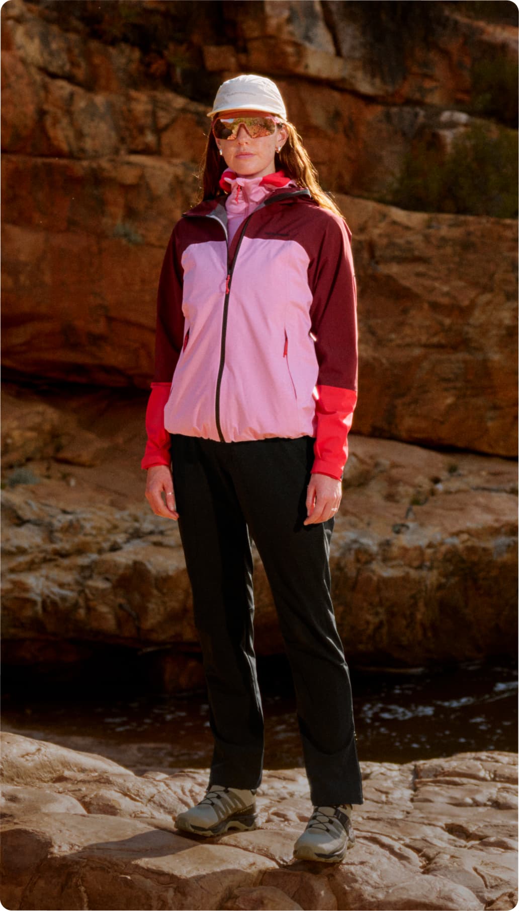 Person in outdoor gear, including a pink and maroon jacket, black pants, and cap, standing on rocky terrain with a cliff backdrop.