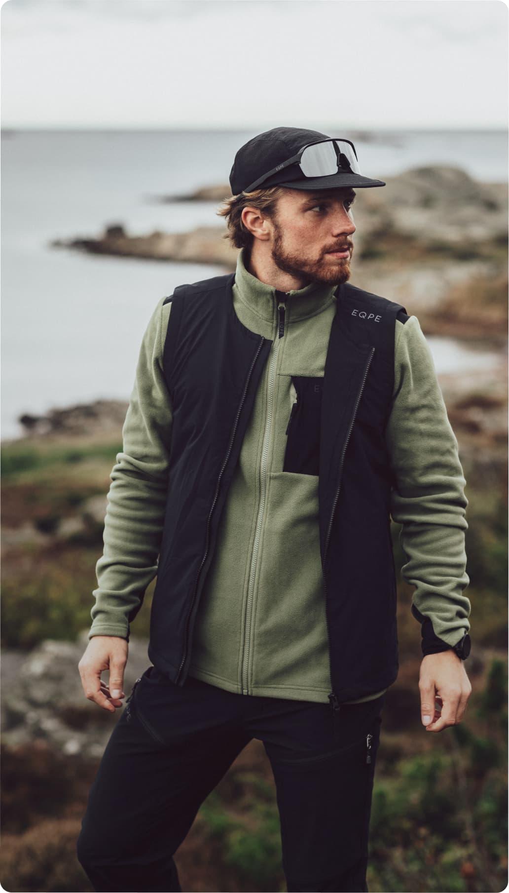 Man in outdoor gear with a green zip-up jacket and black cap stands near a rocky coastline, gazing into the distance.