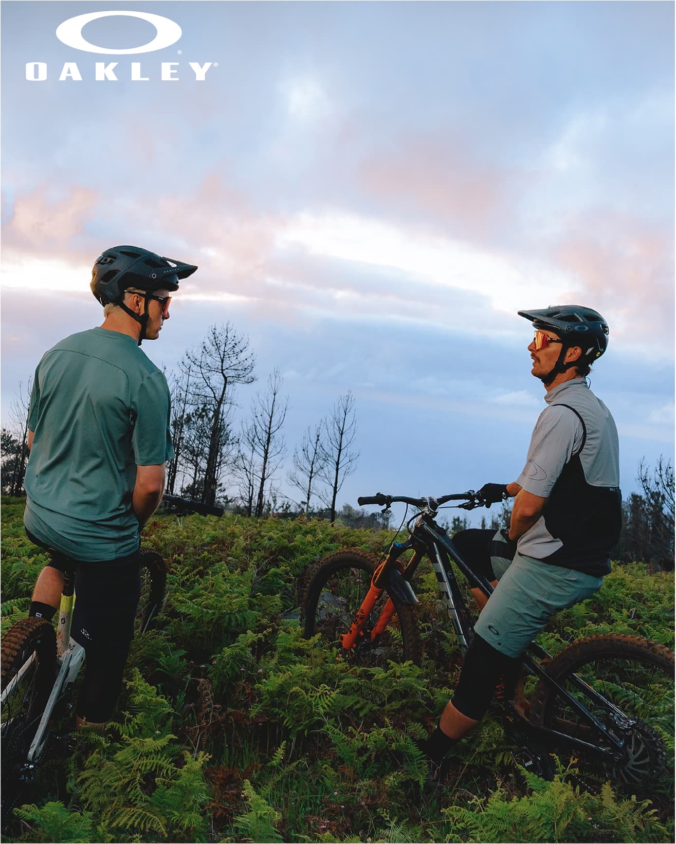 Two cyclists wearing helmets and gear rest beside mountain bikes in a grassy area at sunset, with an Oakley logo above.