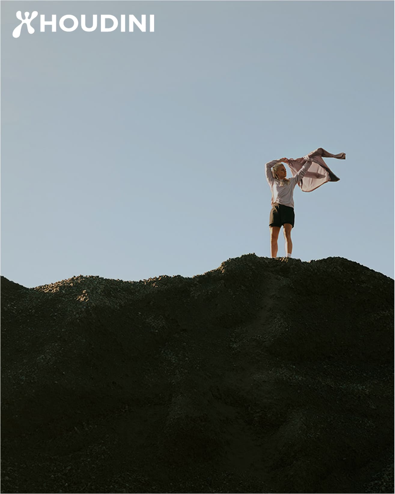 Person standing atop a large hill, holding a jacket against a clear sky. Houdini logo in the top left corner.