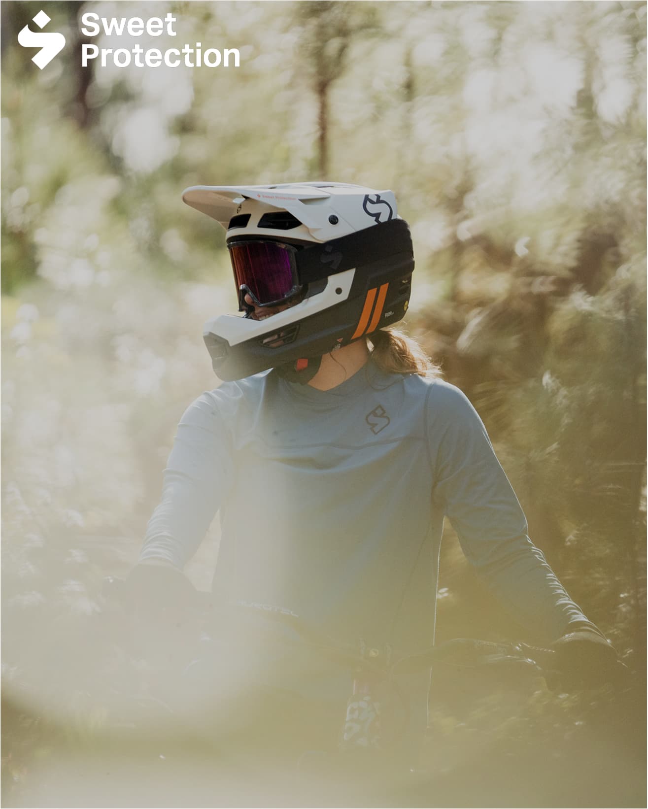 Mountain biker in a white full-face helmet and blue jersey riding through a sunlit forest. Logo reads "Sweet Protection" in the top left corner.