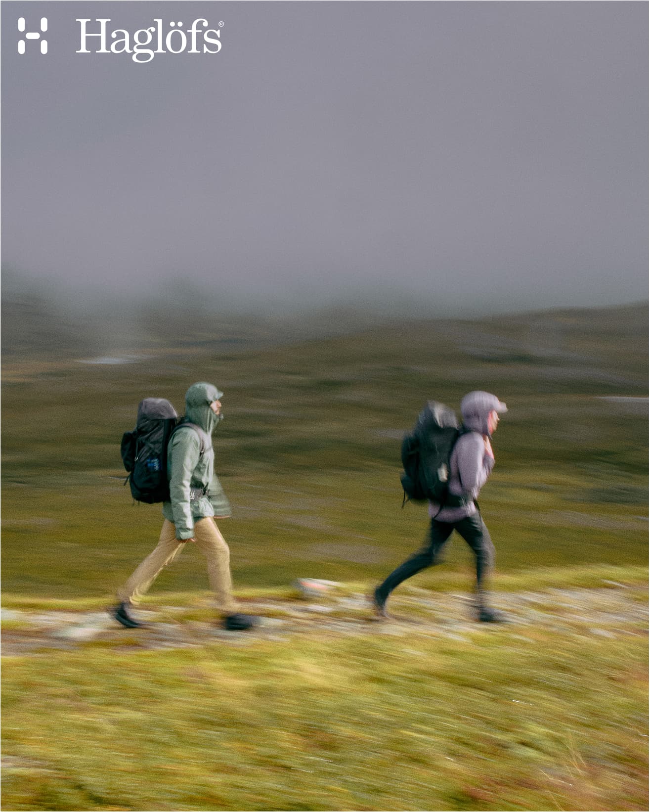 Two hikers wearing jackets and backpacks walk briskly through a foggy landscape, featuring the Haglöfs logo.