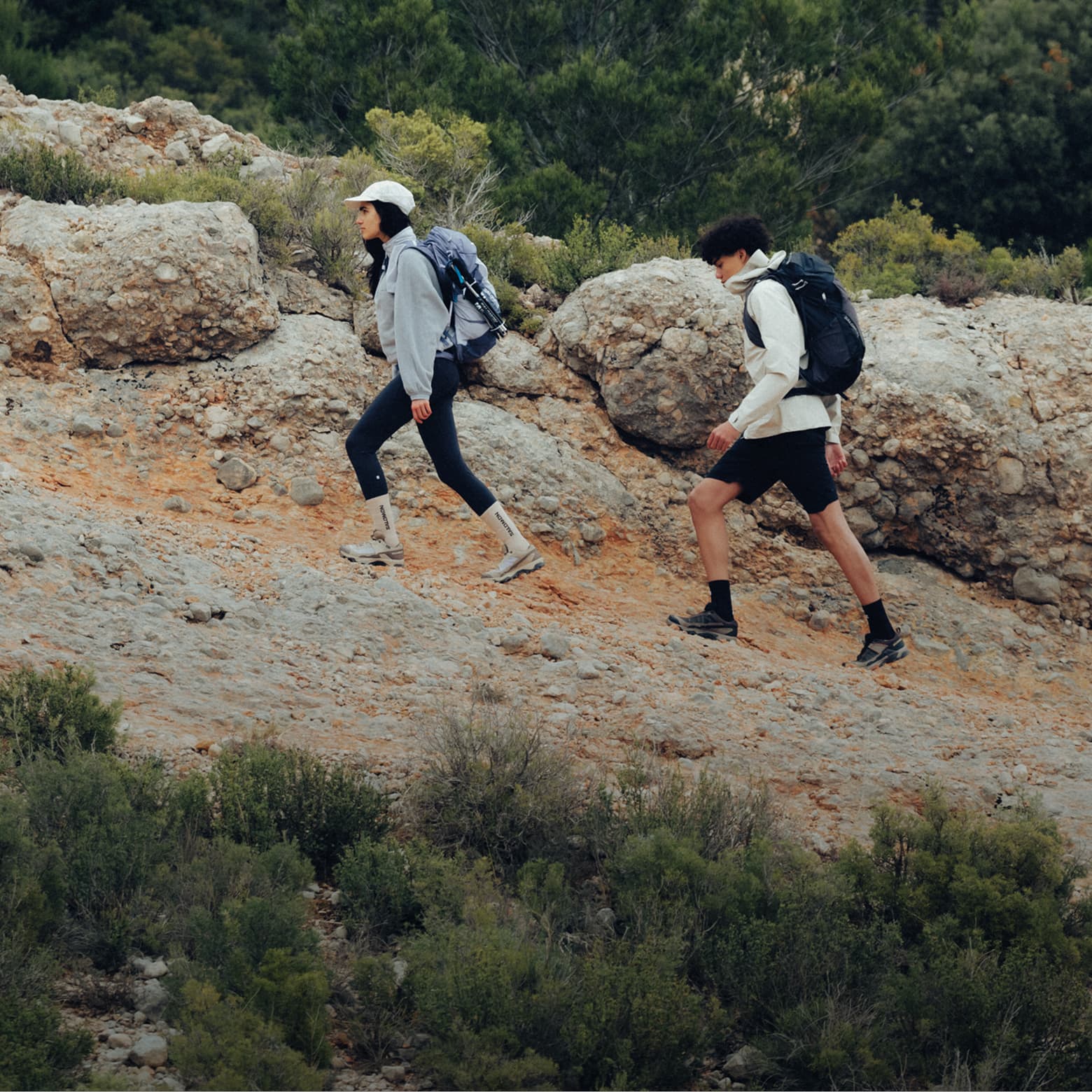 Two hikers with backpacks walk up a rocky trail, surrounded by greenery, wearing casual athletic clothes and hats.