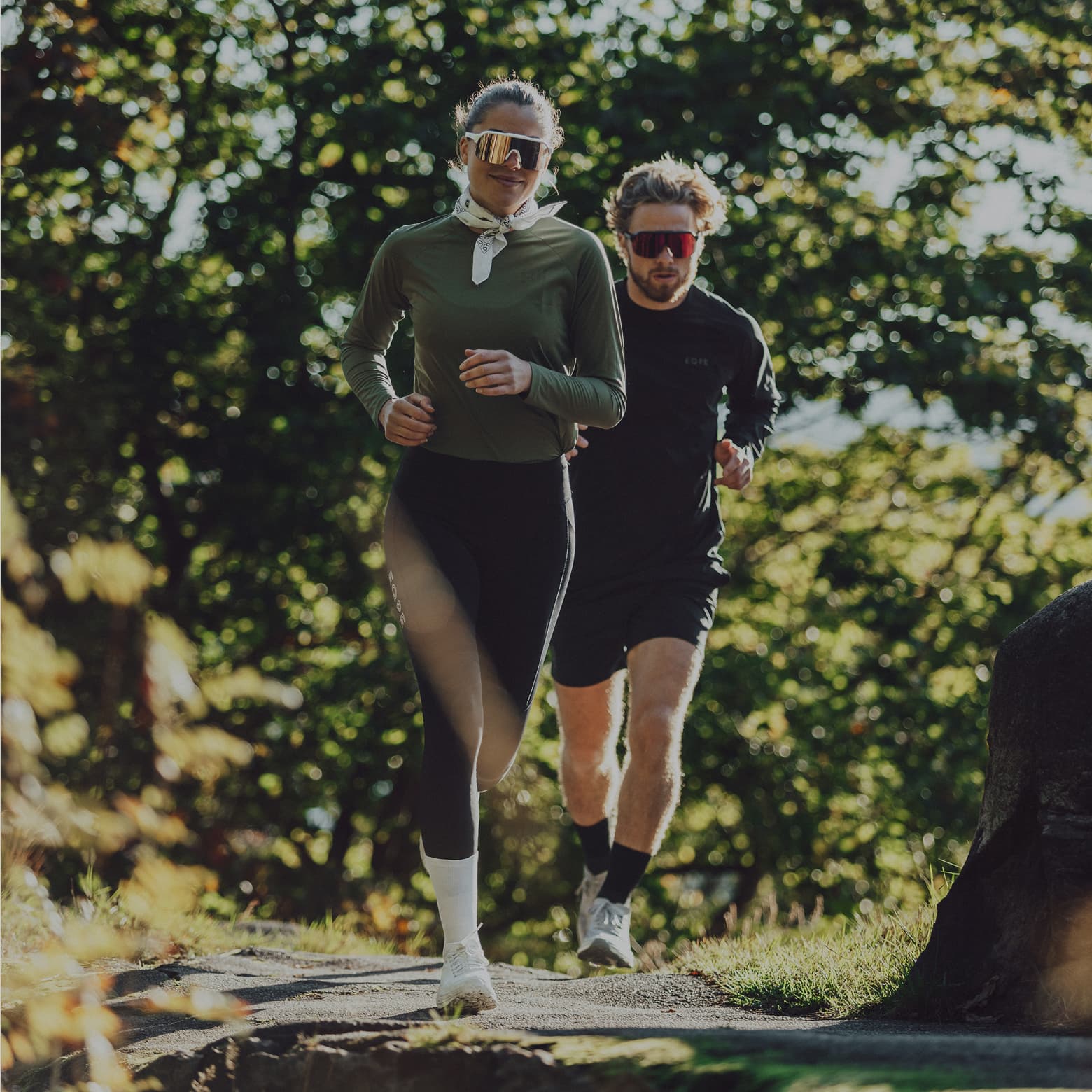 Two people jogging on a forested path, wearing athletic wear and sunglasses, with sunlight filtering through the trees.
