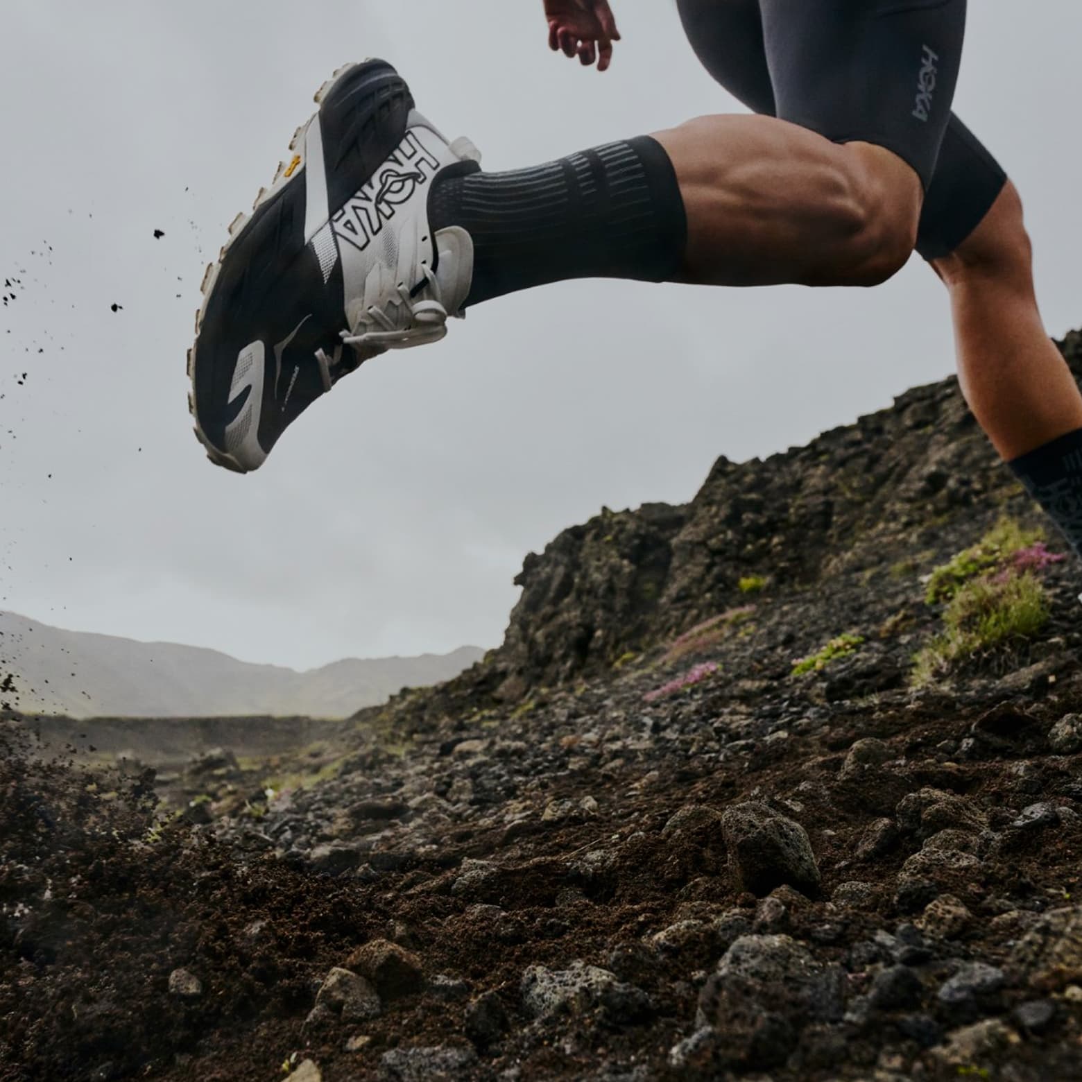 Person running on rocky terrain, kicking up dirt. Wearing black shorts and athletic shoes, emphasizing leg muscles and motion.
