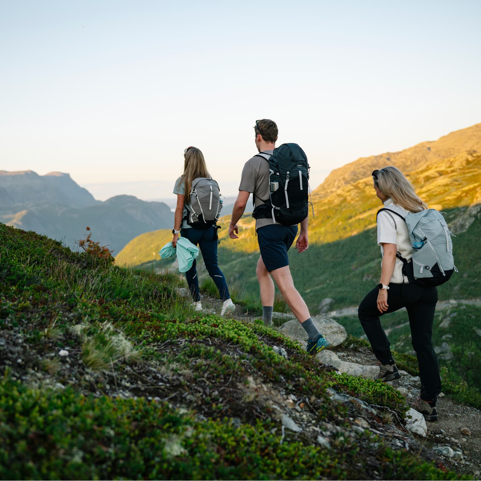 Three hikers with backpacks trekking up a grassy mountain path at sunset, overlooking distant peaks in the background.