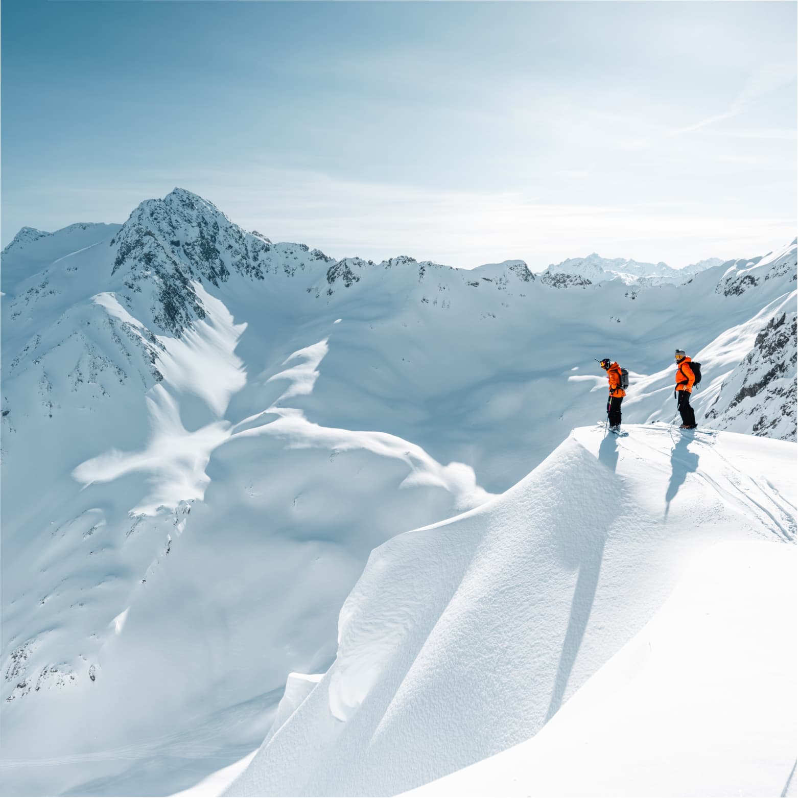 Two skiers in orange jackets stand on a snowy mountain peak, overlooking a vast, snow-covered landscape under a clear blue sky.