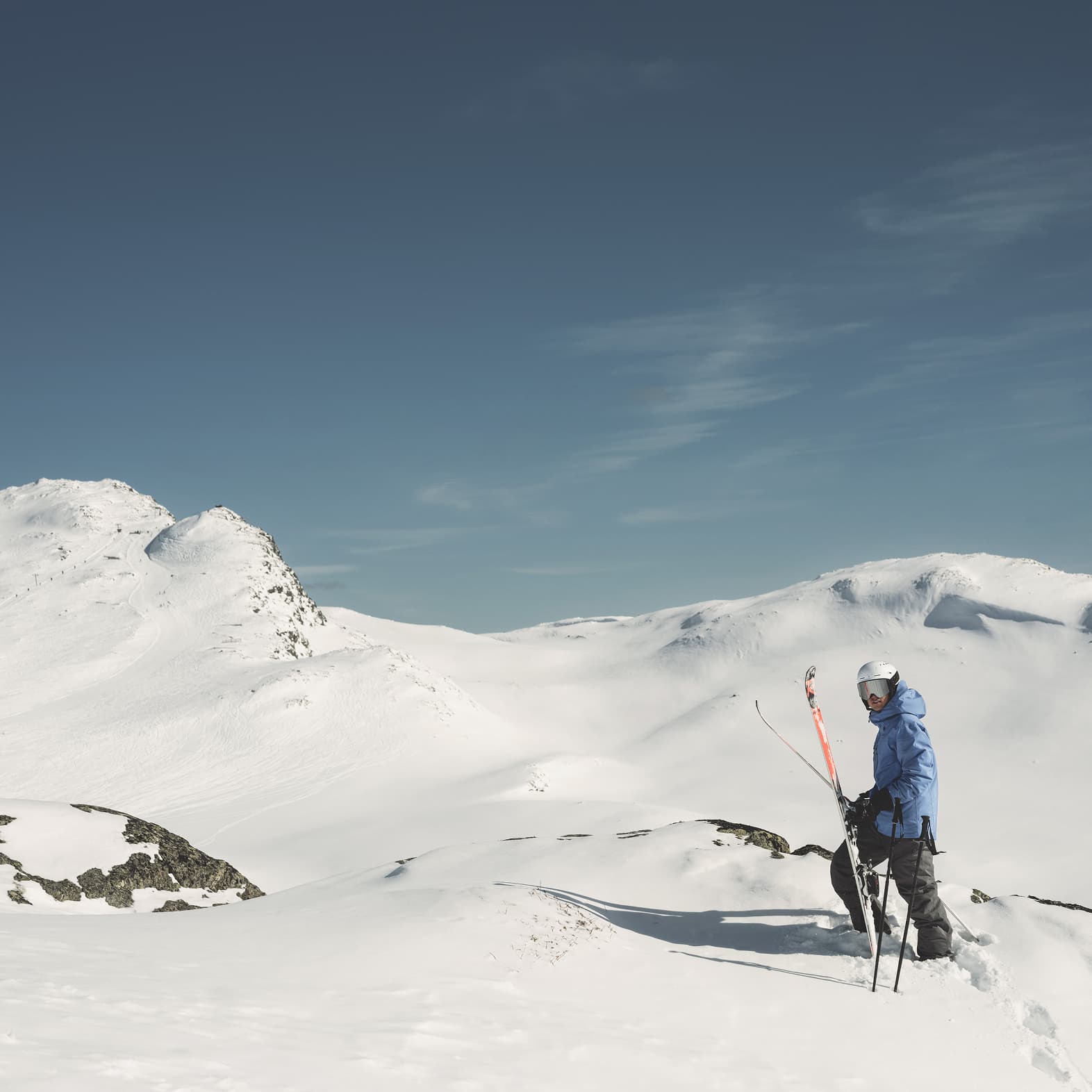 Skier standing on a snowy mountain slope under a clear blue sky, holding ski poles, with distant snow-covered peaks in the background.