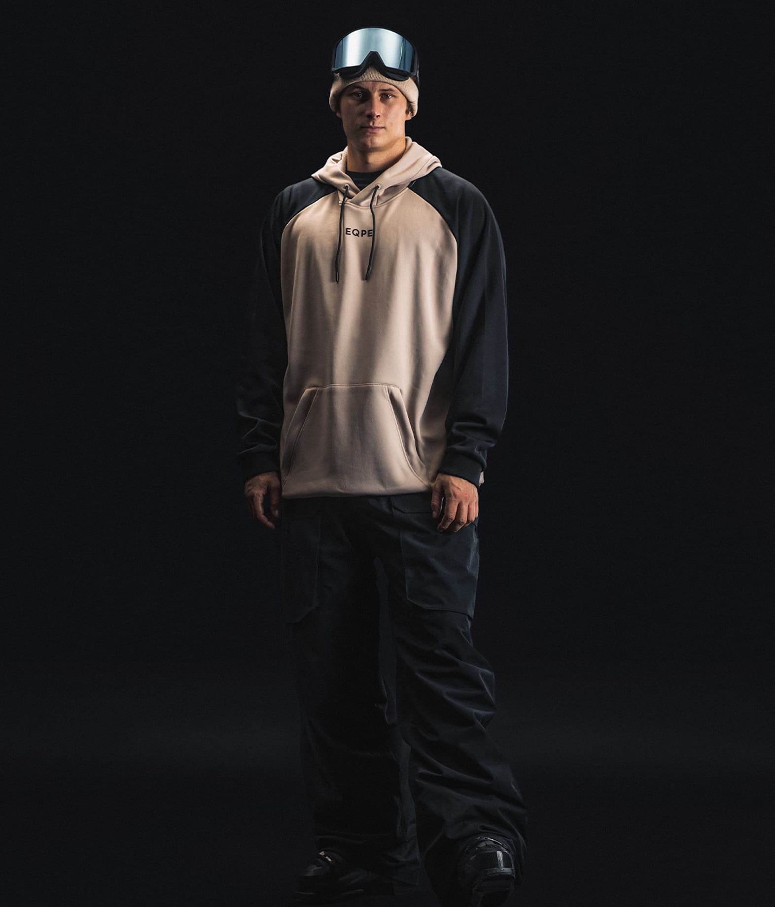A person wearing a hoodie, ski goggles, stands against a dark background.