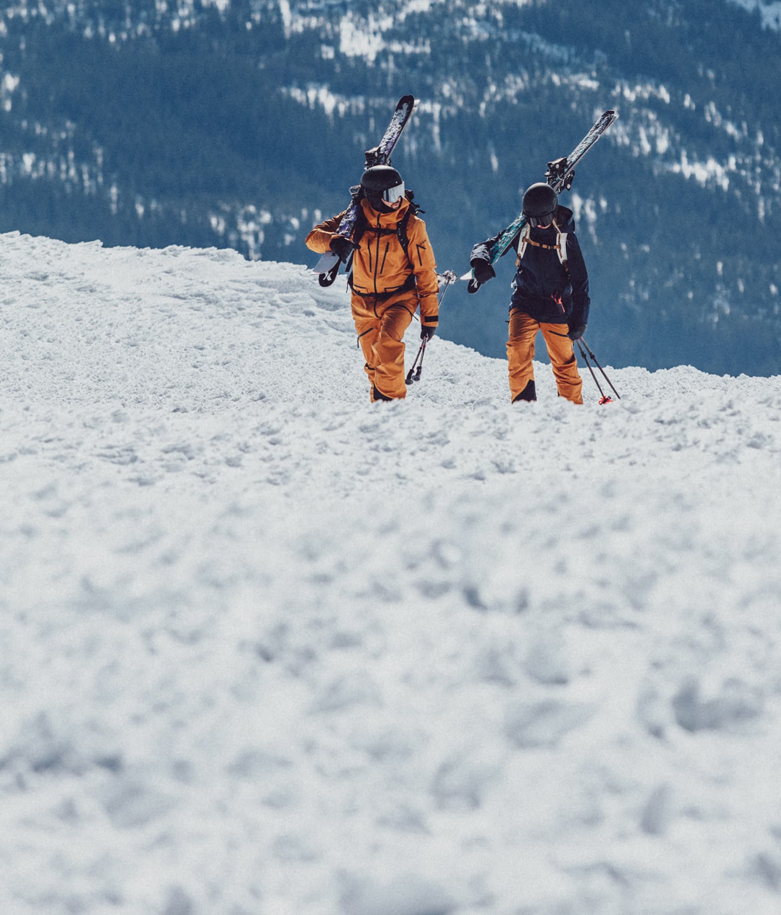 Two skiers carry skis on their shoulders, walking through snowy terrain with a forested mountain backdrop.