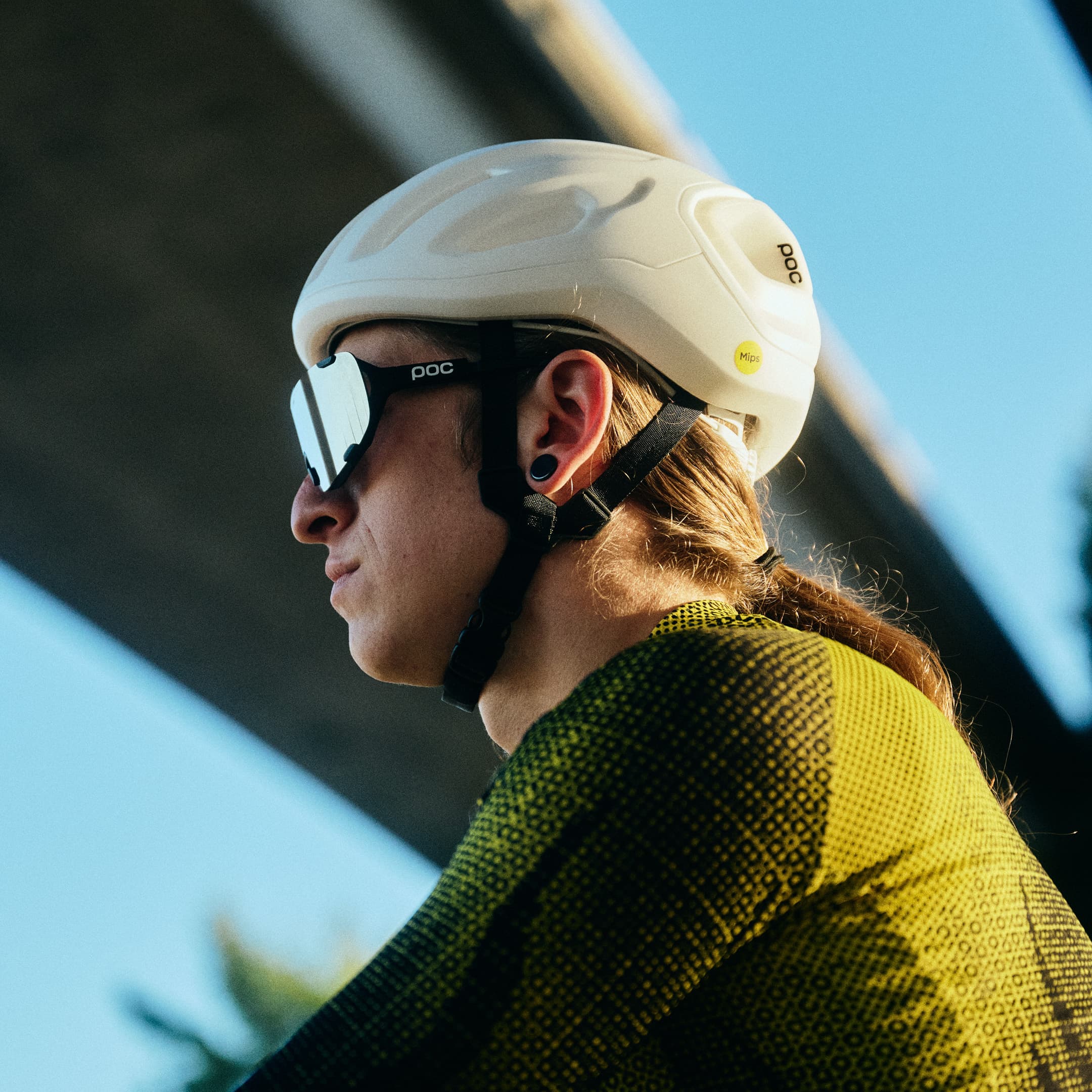 Profile of a cyclist wearing a white helmet and sunglasses, with a textured yellow outfit, under a bridge on a clear day.