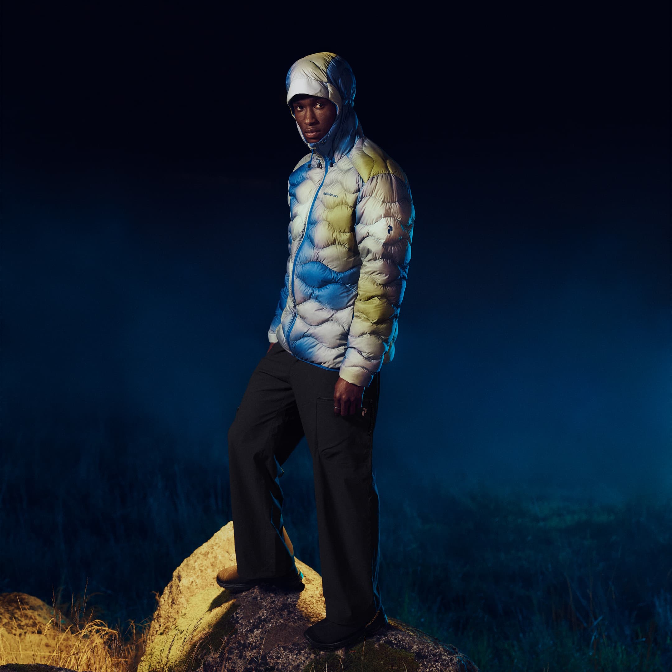 Man in a multicolored puffer jacket stands on a rock against a dark, foggy background at night.